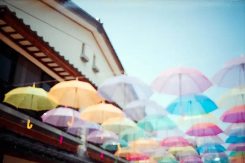 建物間に吊り下げられた色とりどりの傘のインスタレーション。黄色、オレンジ、紫、青、ピンクなど様々な色の傘が空中に浮かび、青空を背景に鮮やかな天蓋を作り出している